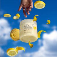 The Clean 2-in-1 Scalp Scrub + Shampoo: Yuzu floating in the air along with scattered slices and peels of yuzu fruit against a backdrop of a bright blue sky with fluffy white clouds. A hand at the top of the image reaches down, outstretched towards the scrub. 