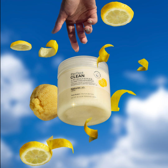 The Clean 2-in-1 Scalp Scrub + Shampoo: Yuzu floating in the air along with scattered slices and peels of yuzu fruit against a backdrop of a bright blue sky with fluffy white clouds. A hand at the top of the image reaches down, outstretched towards the scrub. 