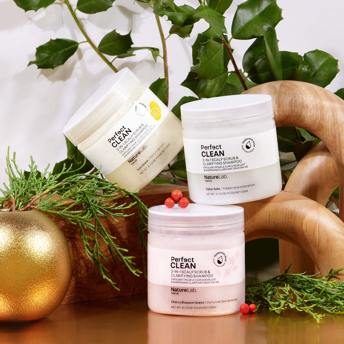 Three jars of the different scents of the Perfect Clean 2-in-1 Scalp Scrub & Clarifying Shampoo, stacked on abstract wooden structures and planters. Holly berries, green plants and branches, and a circular gold planter add a festive touch.