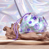 The NatureLab Tokyo x BYBBA limited edition drawstring pouch on a rustic wooden branch in front of a shining metallic background with subtle rainbow reflections. The pouch fabric is a pale green and white checker pattern with Japanese-style flowers in various sizes and shades of purple, and there is also a stamp showing a maneki neko (lucky cat) and the words 'NATURELAB TOKYO' inside, also in a dark purple shade.