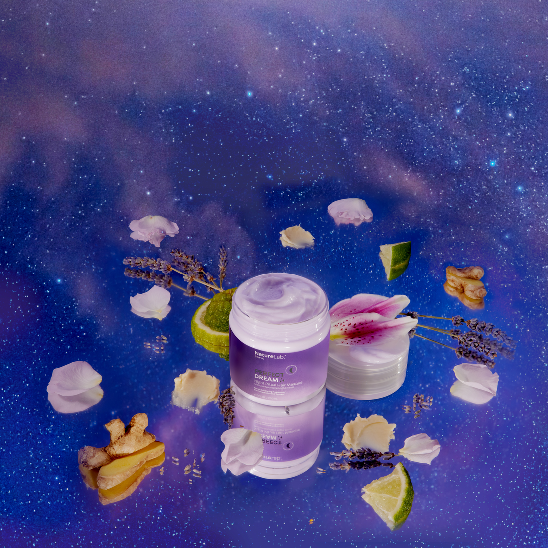 A photo of the Perfect Dream Night Ritual Masque from a high angle, showing much of the top of the jar and the masque's creamy texture. The jar is surrounded by some of its component ingredients: pieces of ginger, flower petals, lavender sprigs, and wedges of fruit. The surface beneath all of these items is a wide expanse of a starry sky pattern, mostly cobalt blue with some faded purple galaxy clouds at the edges. The effect is dreamy and ethereal.